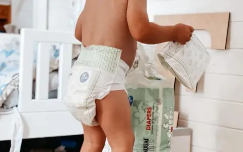 Un bimbo con un pannolino indosso, vicino a una confezione di pannolini. Sembra che i pannolini scadano, ma non c'è fretta.