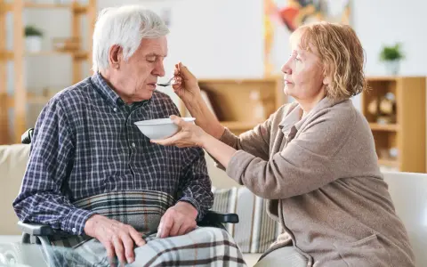 Una donna nutre con un cucchiaino il suo anziano nonno, che non mangia più, seduto su una sedia a rotelle.