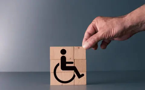 Simboli di accessibilità su cubi di legno, con una mano che aggiunge l'ultimo pezzo. Rappresenta il **certificato medico introduttivo per accompagnamento**.