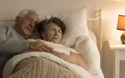 Un uomo anziano abbraccia dolcemente la moglie a letto. La sua mano sulla sua pancia, forse un gesto per confortarla, perché un malato di Alzheimer dorme sempre, cercando riposo.