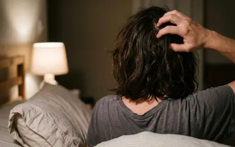 Persona a letto che si gratta la testa, forse perché il prurito aumenta di sera. Luce soffusa da una lampada.