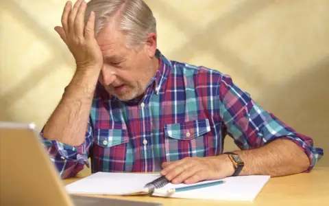 Uomo anziano con mano sulla fronte, frustrato dal non ricordare le cose appena fatte, con quaderno e penna.