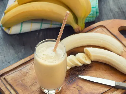 Frullato di banana in un bicchiere con una cannuccia, banane intere e affettate su un tagliere. Con la diarrea si può mangiare la banana, è un alimento delicato.