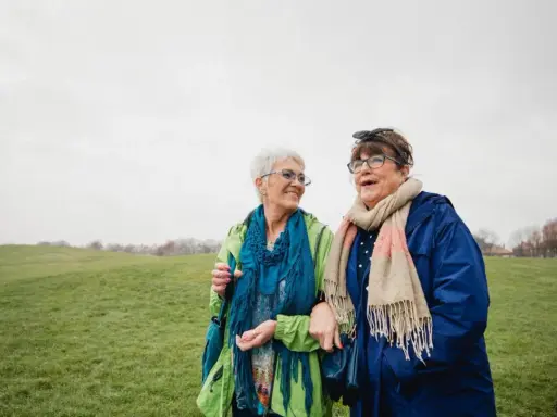 Due anziane passeggiano in un prato. Una delle due, con un foulard blu, sembra avere difficoltà a camminare, ma l'altra le offre supporto, un gesto di cura che va oltre l'Alzheimer.