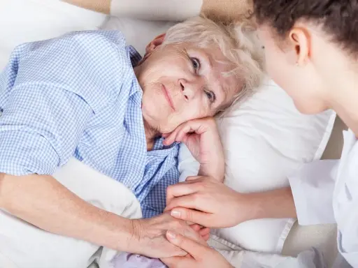 Anziana a letto, assistita da un infermiere. La sua mano stringe quella dell'assistente, forse per alleviare il dolore di una piaga da decubito sull'osso sacro.
