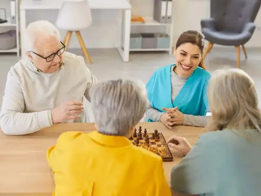 Un gruppo di anziani gioca a scacchi, un'attività che promuove la stimolazione cognitiva. Un'assistente sorride osservando.