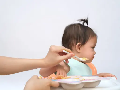 Bambina seduta sul seggiolone, con un cucchiaino di cibo offerto da una mano. Un modo per stimolare l'appetito con pazienza e amore.