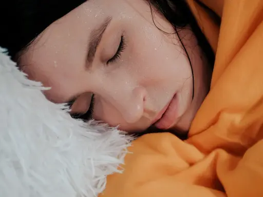 Viso di donna con gocce di sudore sulla fronte, avvolta in una coperta arancione e piumino bianco. Sembra che stia per sudare freddo.
