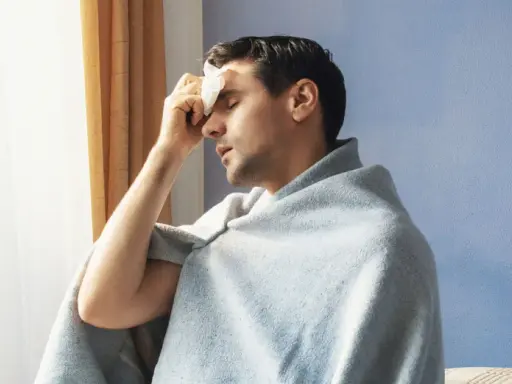 Uomo con panno umido sulla fronte, avvolto in una coperta, che sembra soffrire di sudori freddi.