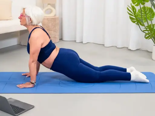 Donna anziana in posa yoga cobra su tappetino blu, seguendo un tutorial online. Un esempio di palestra per anziani attiva e moderna.