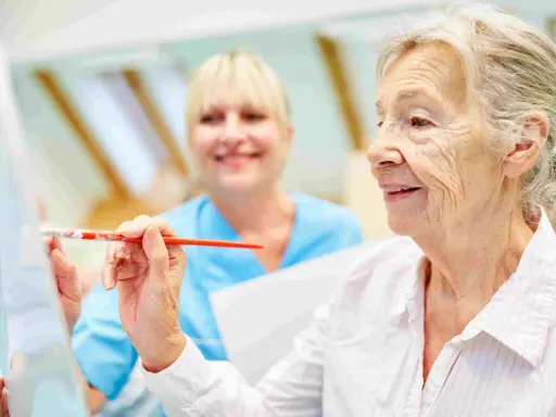 Anziana dipinge con un pennello, assistita da un'infermiera sorridente. Un esempio di terapie non farmacologiche per l'Alzheimer che stimolano la creatività.