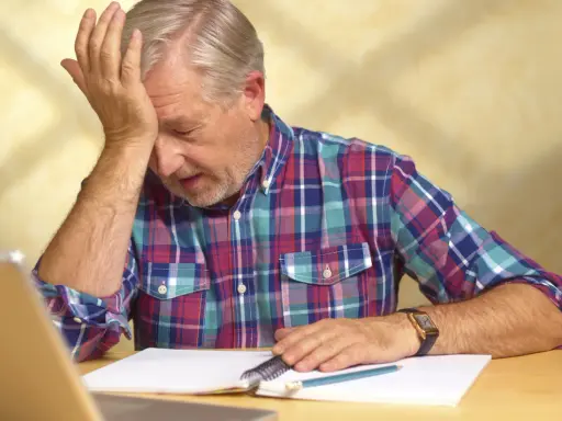 Uomo anziano con mano sulla fronte, frustrato dal non ricordare le cose appena fatte, con quaderno e penna.
