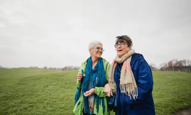 Due anziane passeggiano in un prato. Una delle due, con un foulard blu, sembra avere difficoltà a camminare, ma l'altra le offre supporto, un gesto di cura che va oltre l'Alzheimer.