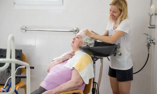 Assistente che lava i capelli a un'anziana non autosufficiente in un bagno attrezzato, con cura e attenzione.