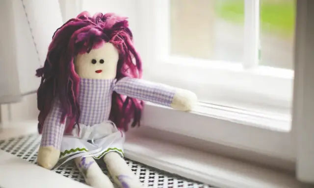 Bambola con capelli viola, vestita a quadretti, seduta sul davanzale. Un simbolo di conforto nella doll therapy per l'Alzheimer.