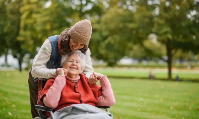 Una giovane donna sorride mentre abbraccia un'anziana in sedia a rotelle in un parco. Un momento di gioia che mostra come un caregiver può sopravvivere con amore.