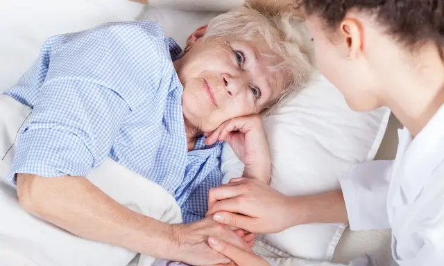 Anziana a letto, assistita da un infermiere. La sua mano stringe quella dell'assistente, forse per alleviare il dolore di una piaga da decubito sull'osso sacro.