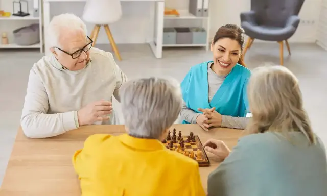 Un gruppo di anziani gioca a scacchi, un'attività che promuove la stimolazione cognitiva. Un'assistente sorride osservando.