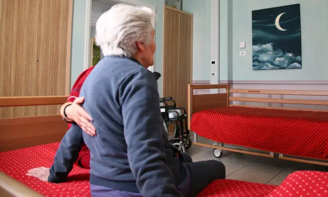 Anziana in stanza, forse affetta da sindrome da ospedalizzazione, con un'altra persona che le appoggia una mano sulla spalla. Sulla parete un quadro con luna e nuvole.