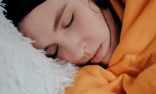 Viso di donna con gocce di sudore sulla fronte, avvolta in una coperta arancione e piumino bianco. Sembra che stia per sudare freddo.