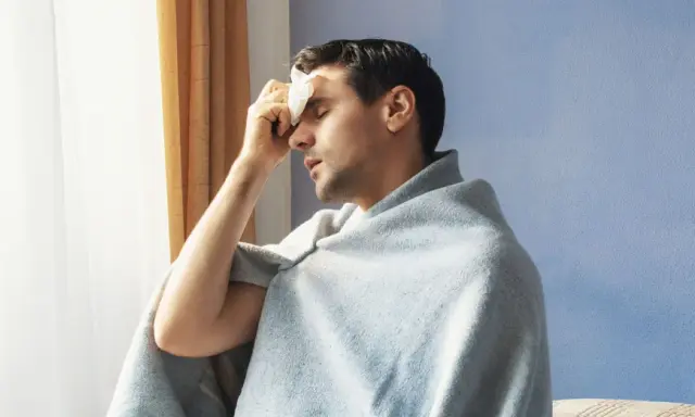 Uomo con panno umido sulla fronte, avvolto in una coperta, che sembra soffrire di sudori freddi.