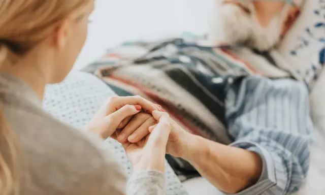 Mani che si stringono tra una giovane donna e un anziano a letto, con cannule nasali. Il caregiver viene pagato per la sua dedizione.