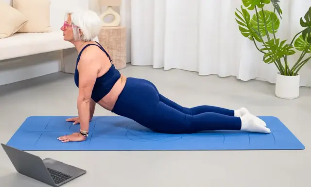Donna anziana in posa yoga cobra su tappetino blu, seguendo un tutorial online. Un esempio di palestra per anziani attiva e moderna.