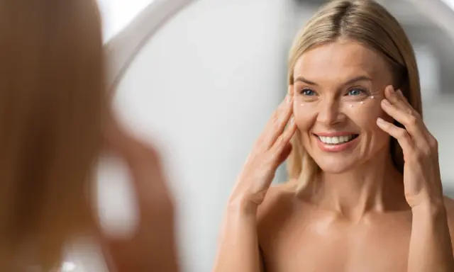 Donna sorride applicando crema contorno occhi. La sua pelle sottile del viso mostra segni del tempo, ma è luminosa.