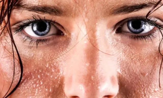 Occhi intensi e pelle lucida di sudore, con goccioline che riflettono la luce. Le vampate di sudore sono evidenti sul viso.