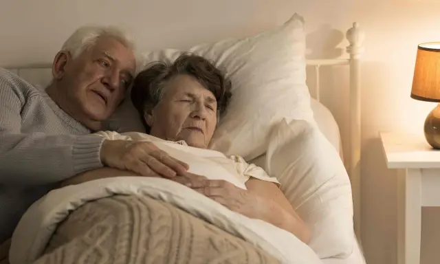 Un uomo anziano abbraccia dolcemente la moglie a letto. La sua mano sulla sua pancia, forse un gesto per confortarla, perché un malato di Alzheimer dorme sempre, cercando riposo.