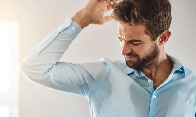 Uomo con camicia azzurra macchiata di sudore sotto le ascelle, che si gratta la testa. Sudare tanto è sintomo di stress o caldo.