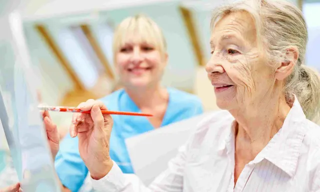 Anziana dipinge con un pennello, assistita da un'infermiera sorridente. Un esempio di terapie non farmacologiche per l'Alzheimer che stimolano la creatività.