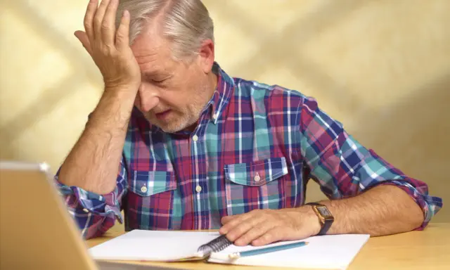 Uomo anziano con mano sulla fronte, frustrato dal non ricordare le cose appena fatte, con quaderno e penna.