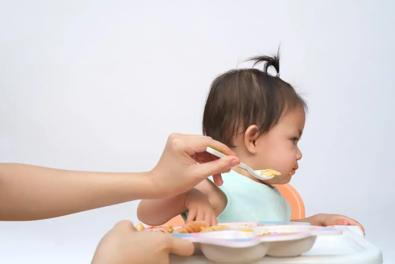 Bambina seduta sul seggiolone, con un cucchiaino di cibo offerto da una mano. Un modo per stimolare l'appetito con pazienza e amore.