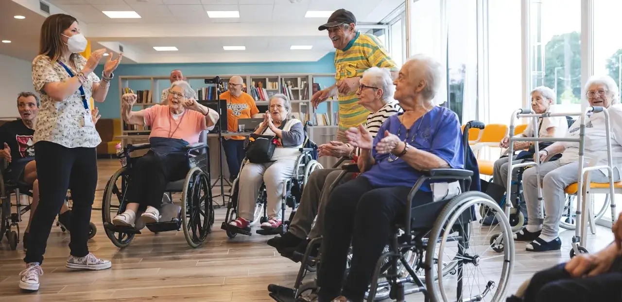 Un gruppo di anziani in sedie a rotelle partecipa a un'attività in una struttura convenzionata per malati di Alzheimer, guidati da un'animatrice.