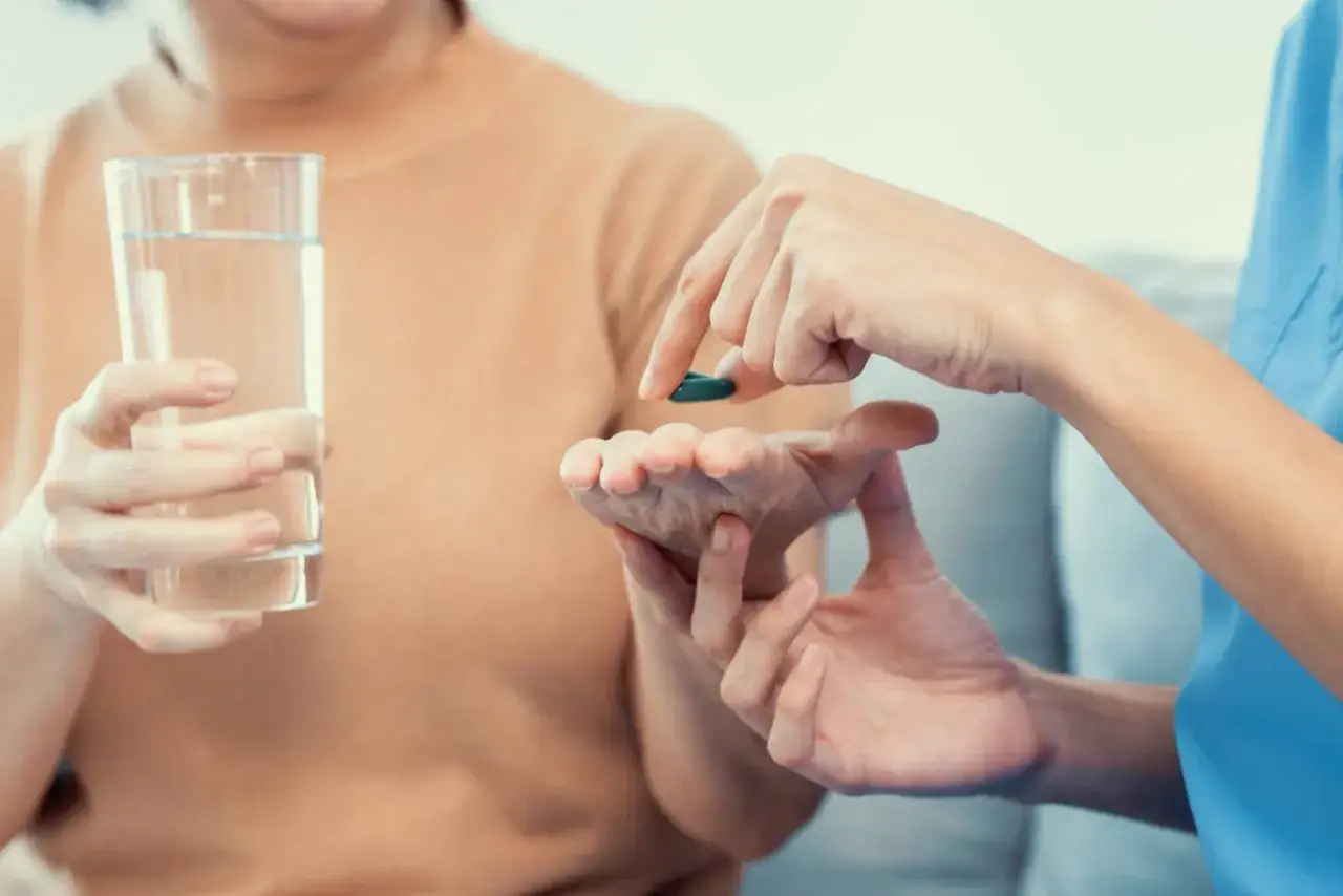 Un assistente socio sanitario porge una pillola a un anziano, che tiene un bicchiere d'acqua.