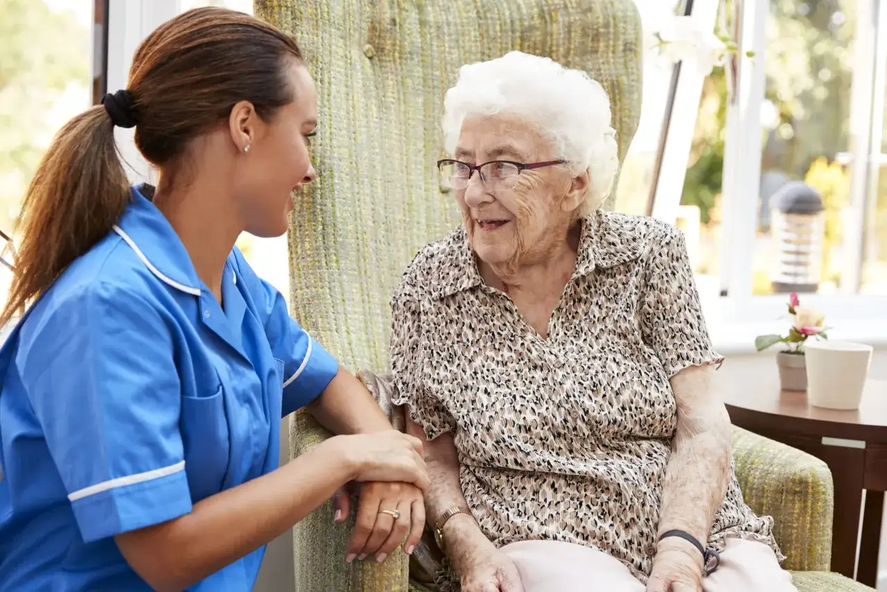Assistente in camice blu parla con un'anziana signora con occhiali, forse mostrando i sintomi finali dell'Alzheimer, in una stanza luminosa.