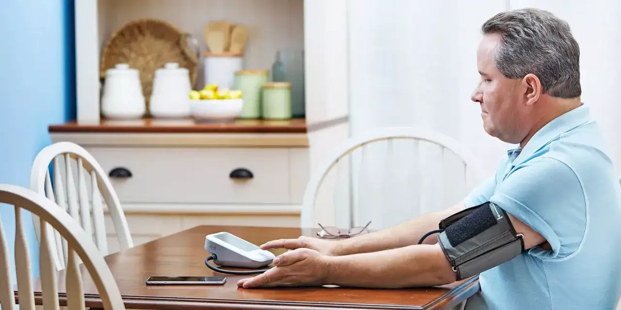 Uomo misura la pressione, preoccupato per l'ipertensione emotiva. Sul tavolo, un misuratore, occhiali e smartphone.