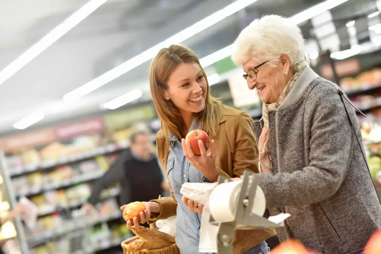 Giovane donna aiuta anziana a scegliere mele al supermercato, un momento di cura che può essere importante per chi manifesta i primi sintomas de alzheimer.