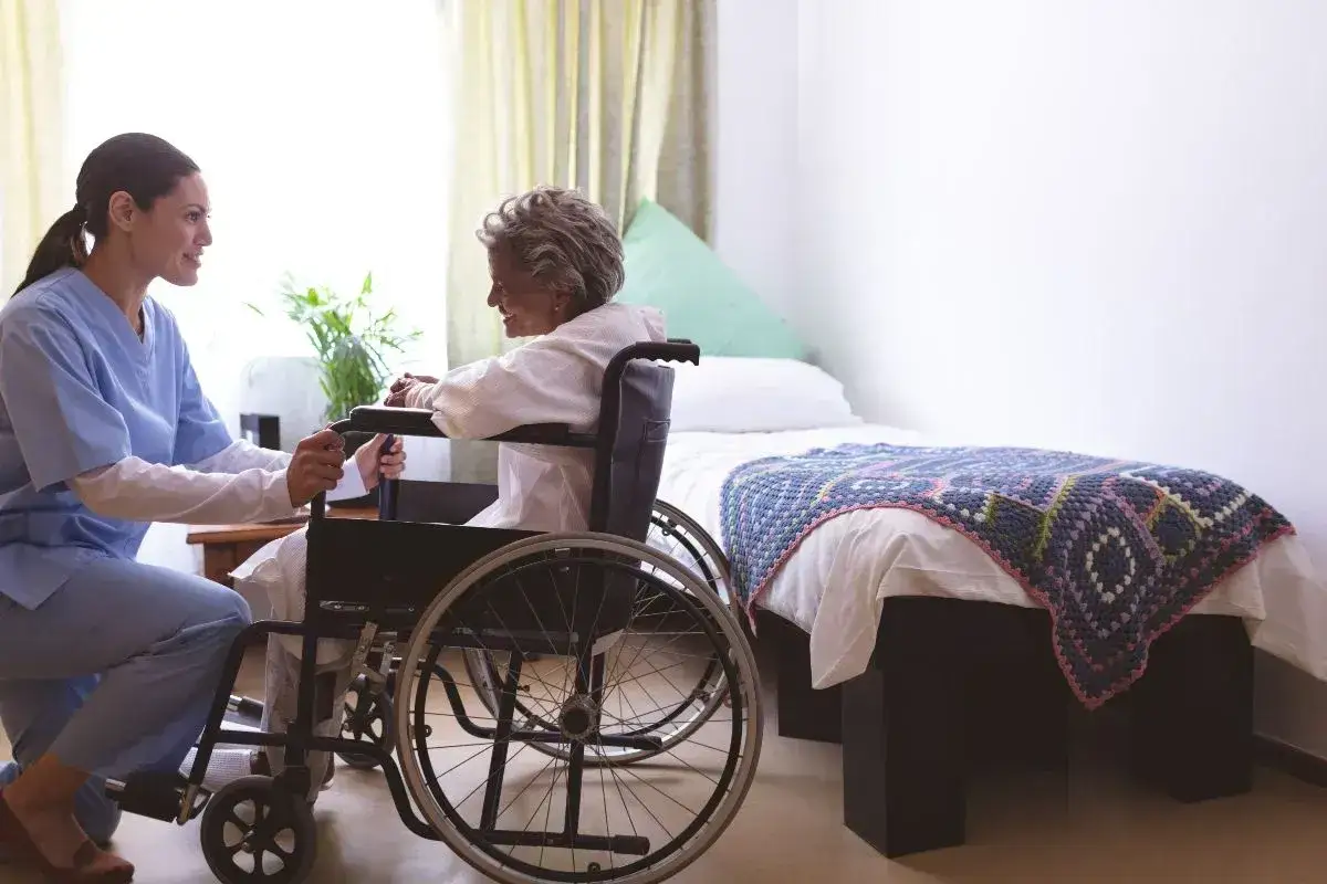 Un caregiver, con un sorriso, assiste un'anziana in sedia a rotelle in una stanza. Il suo ruolo è fondamentale per il benessere.