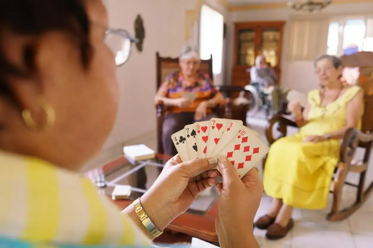Anziani giocano a carte, un'attività che favorisce la stimolazione cognitiva. In primo piano, mani che tengono carte da gioco. Sullo sfondo, altre persone anziane sedute.