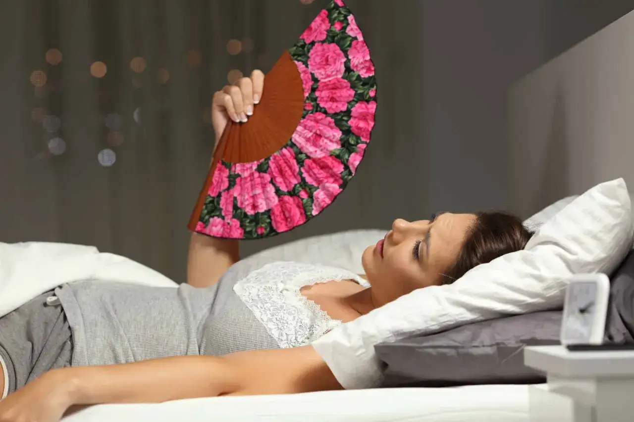 Donna a letto, con un ventaglio fiorato, soffre il caldo. Possibili cause di sudorazione notturna nelle donne.
