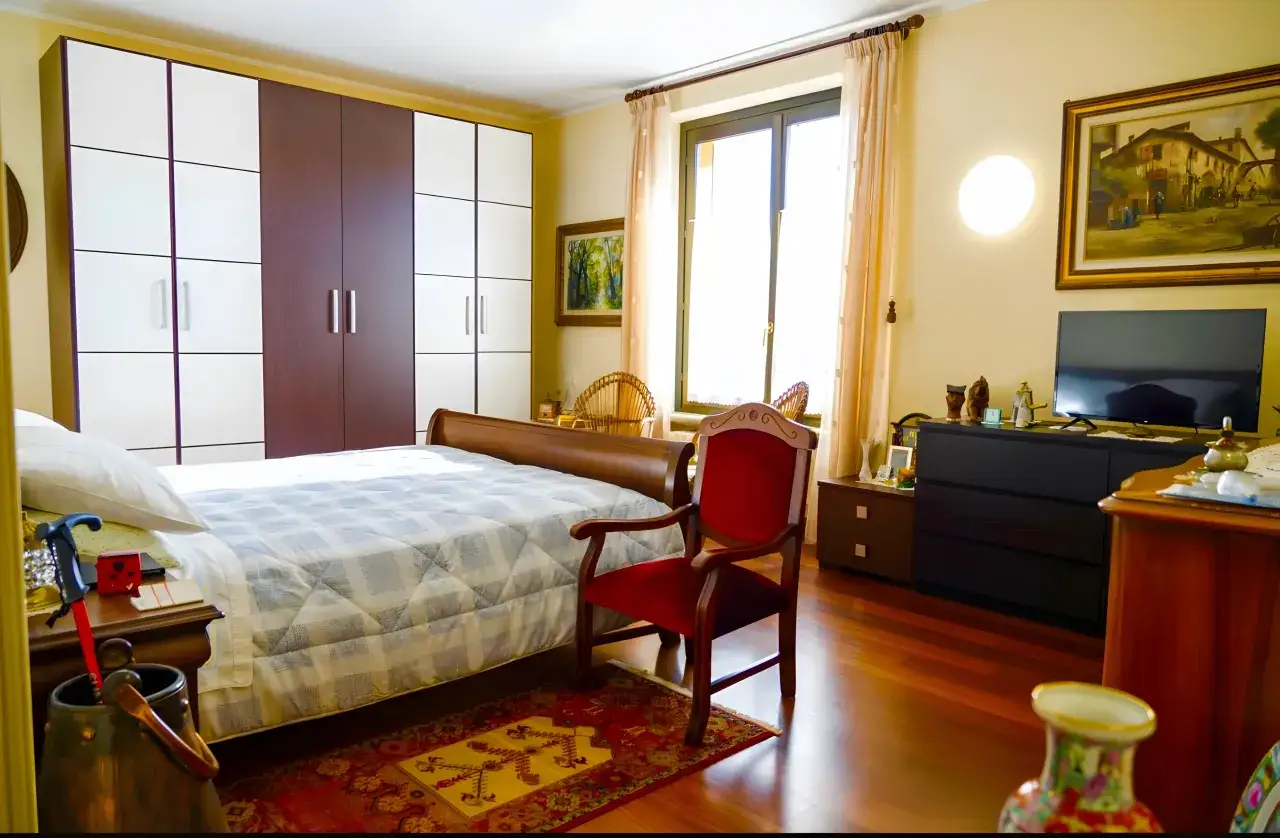 Camera da letto accogliente in una casa famiglia per anziani non autosufficienti, con letto comodo, armadio spazioso e poltrona rossa.