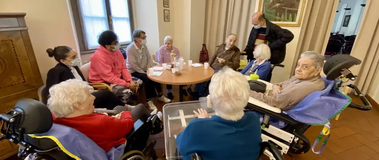 Un gruppo di persone, anziani e operatori, si riunisce attorno a un tavolo in una RSA. Si discute e si socializza, mostrando cosa sono le RSA: luoghi di cura e comunità.