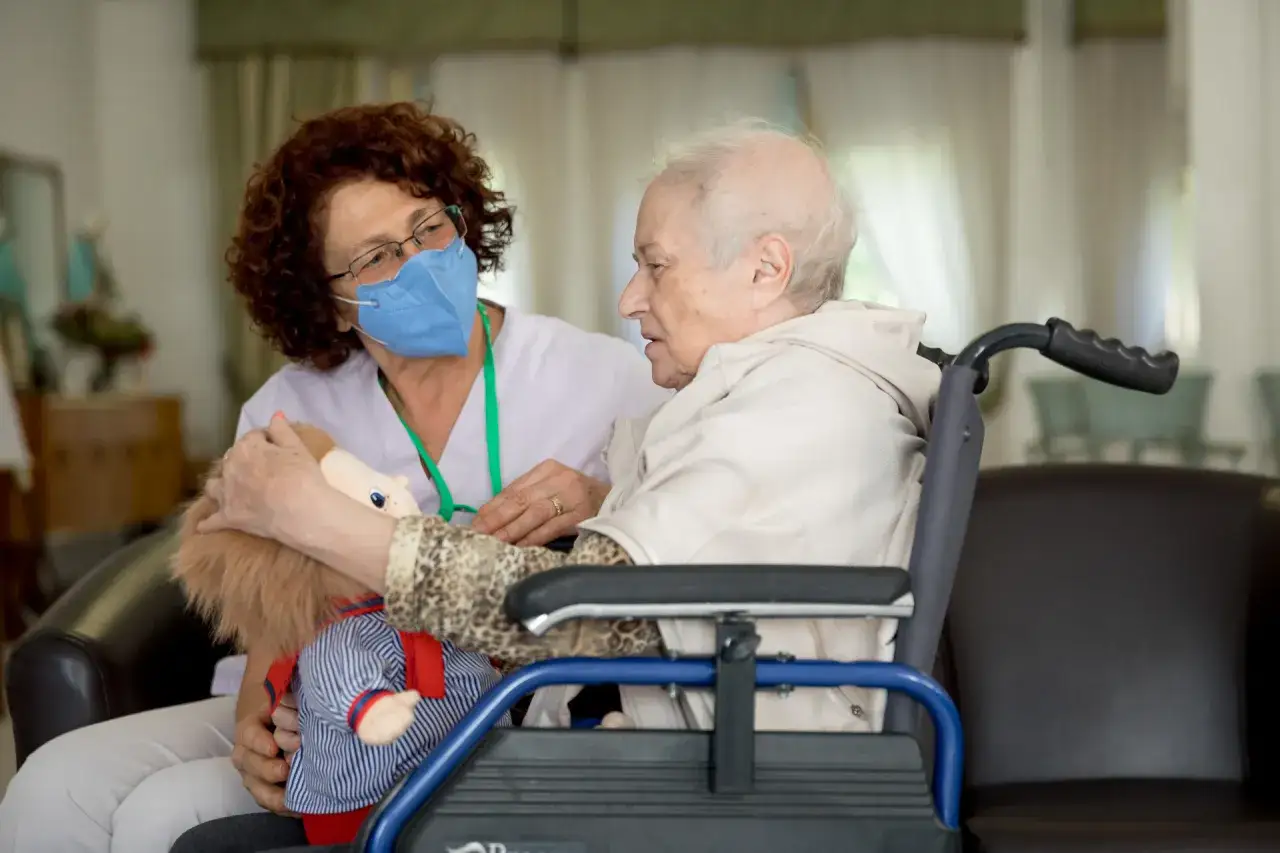 Un'operatrice con mascherina blu mostra una bambola a un'anziana in carrozzina, un esempio di doll therapy per l'Alzheimer.