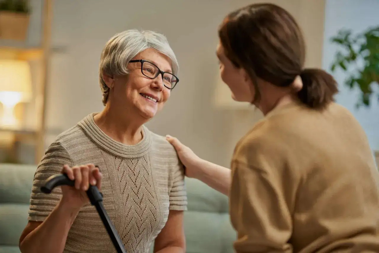 Una donna accarezza la spalla di un'anziana sorridente con un bastone. La demenza senile può influenzare la defecazione, ma l'affetto è un supporto importante.