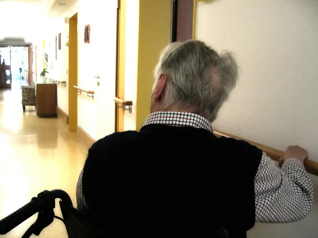 Anziano in sedia a rotelle in corridoio, forse in attesa per la domanda invalidità anziani.