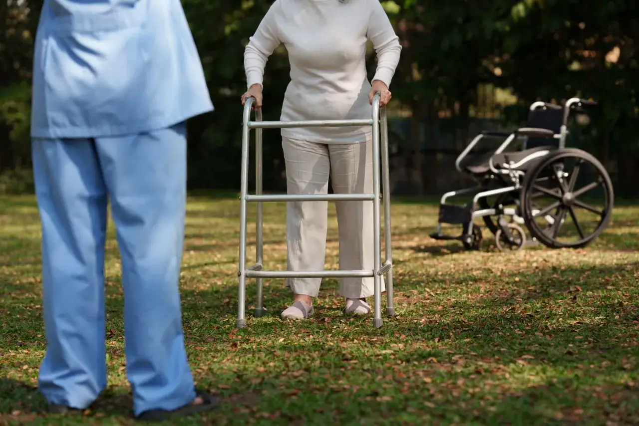 Assistente supporta una persona con alzheimer e difficoltà a camminare, che usa un deambulatore. Sfondo con sedia a rotelle.