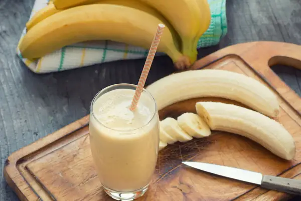 Frullato di banana in un bicchiere con una cannuccia, banane intere e affettate su un tagliere. Con la diarrea si può mangiare la banana, è un alimento delicato.