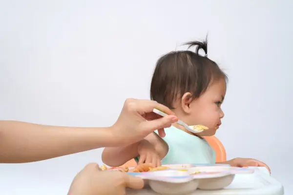 Bambina seduta sul seggiolone, con un cucchiaino di cibo offerto da una mano. Un modo per stimolare l'appetito con pazienza e amore.